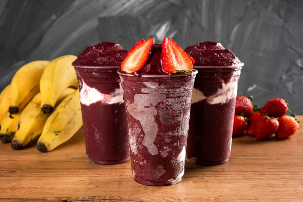 Three Brazilian Frozen Açai Berry Ice Cream Smoothie in plastic cup with Strawberries and condensed milk. On wooden desk and a gray summer background with fruits. Front view for menu and social media Three Brazilian Frozen Açai Berry Ice Cream Smoothie in plastic cup with Strawberries and condensed milk. On wooden desk and a gray summer background with fruits. Front view for menu and social media